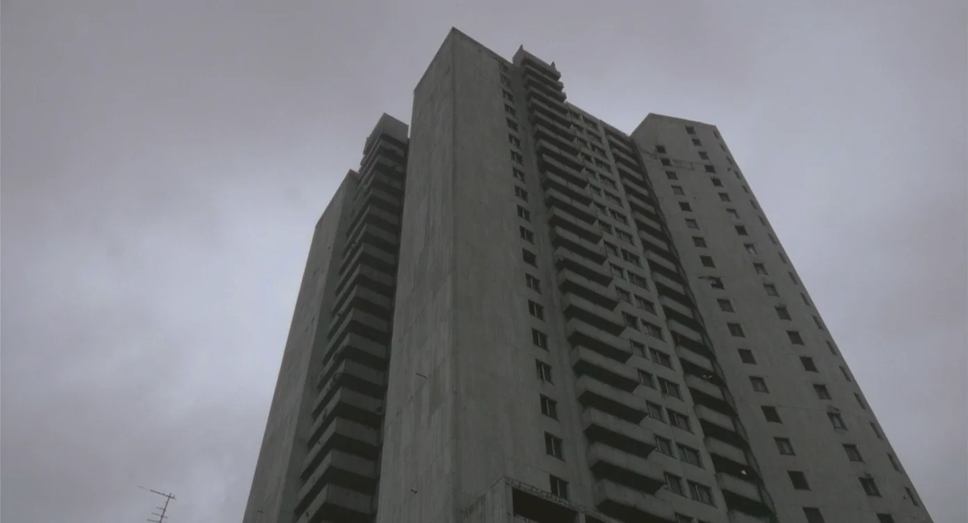 A 1960s Brutalist concrete tower block of flats with a separate service tower, rising against a grey overcast sky