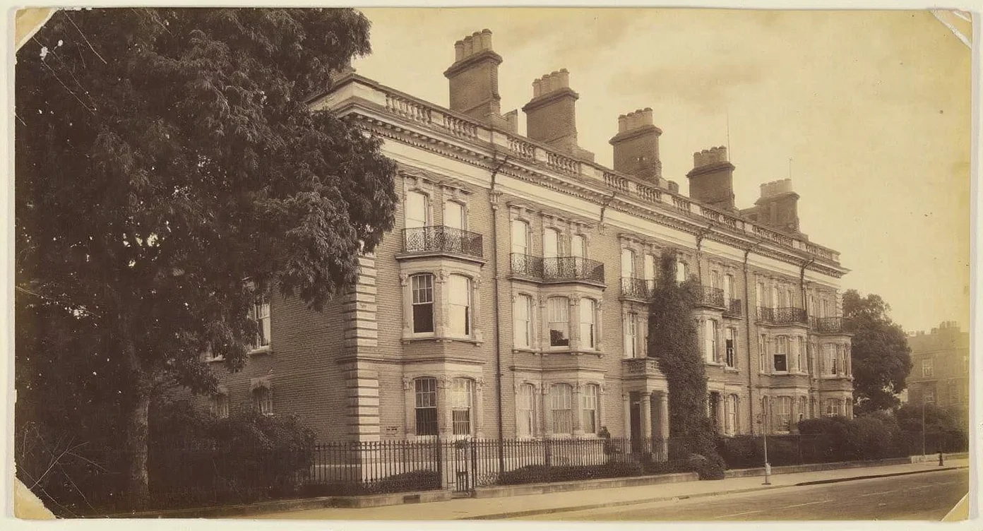 An aged, sepia-toned antique photograph of an early 20th-century London mansion block of flats on a railed residential street with a plane tree