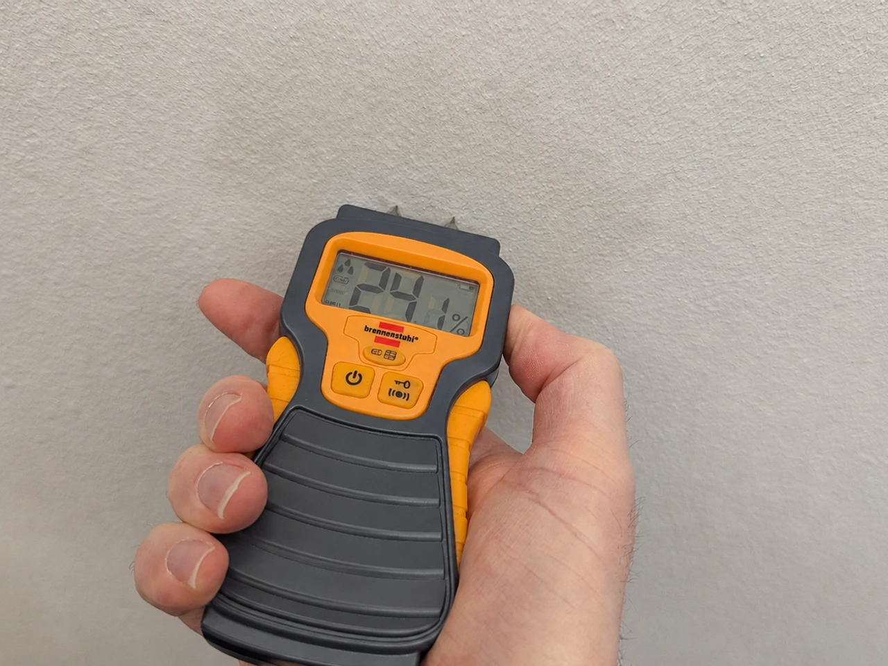 Close-up of a handheld damp meter being held against a painted plaster wall in a UK leasehold flat, the probes pressed into a faint moisture mark.