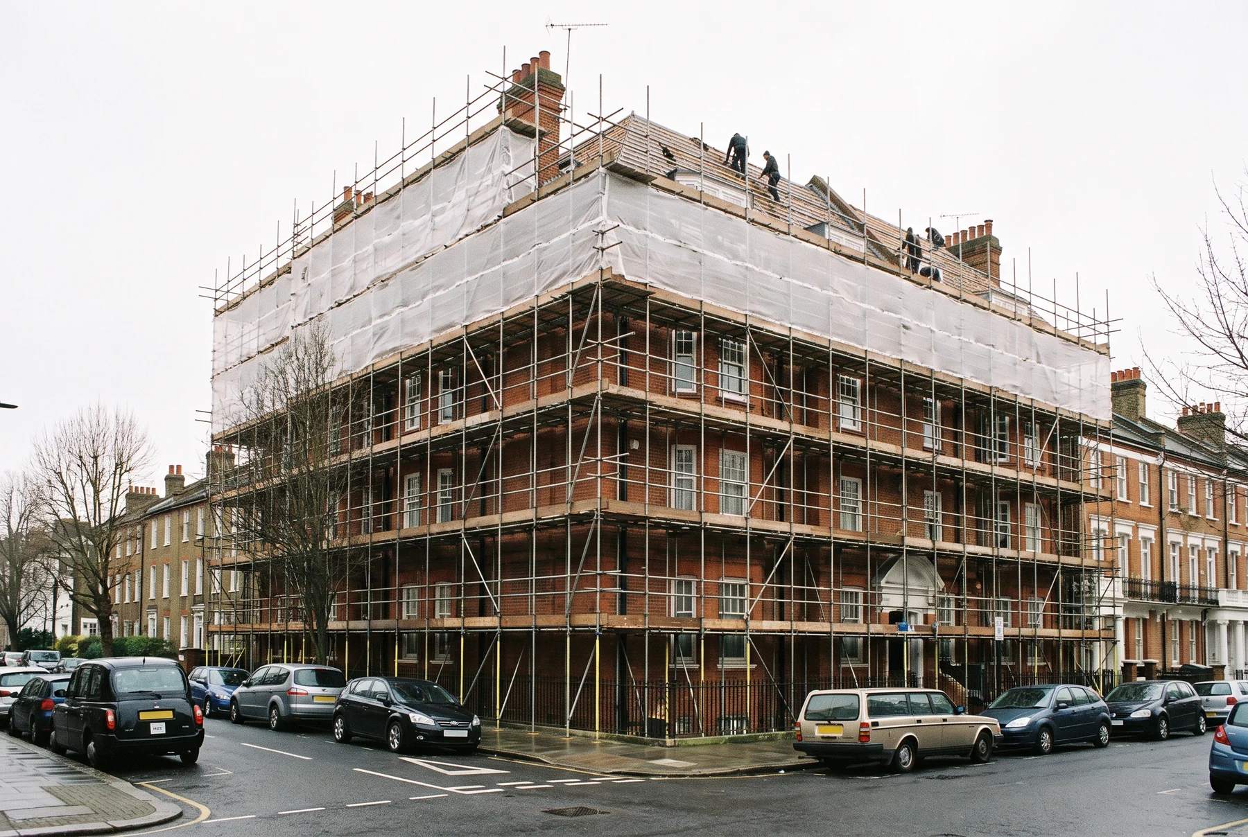 A red-brick mansion block of flats on a London street corner wrapped in scaffolding and white netting, with roofers working on the roof