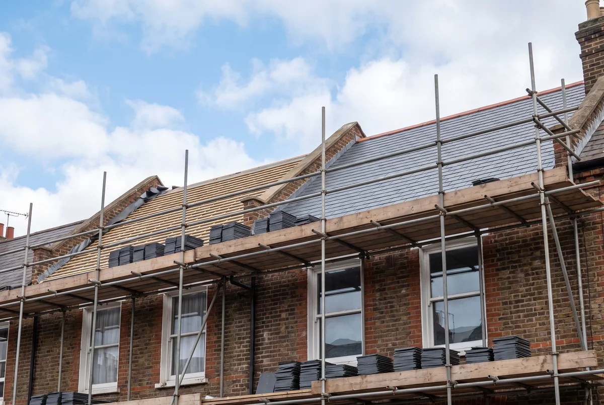 Scaffolding across the front of a terrace of flats while the roof is re-slated, with stacks of new slates on the boards outside the top-floor windows