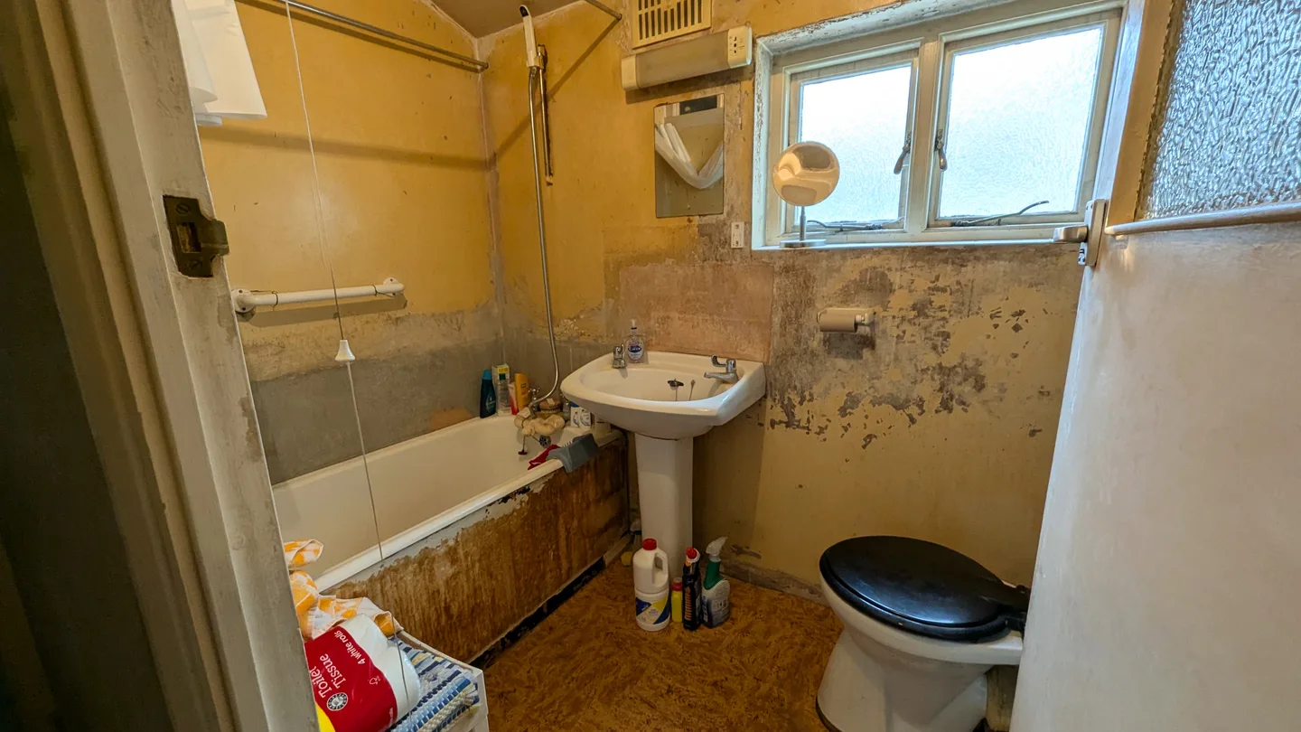 A bathroom in poor condition, with stripped walls, missing tiles and an exposed subfloor