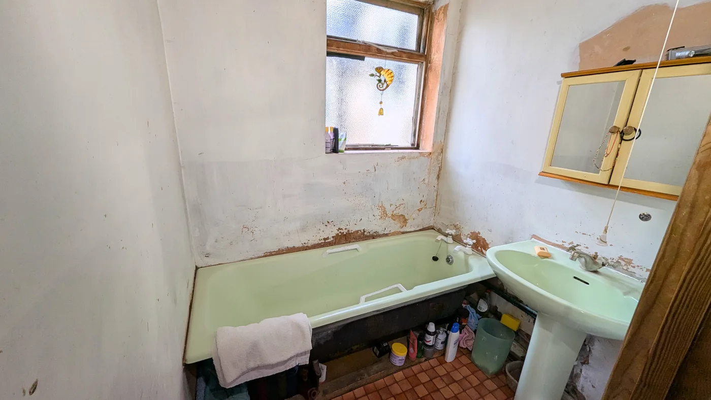 An old bathroom with a pale green bath and basin, damaged plaster and a bare tiled floor