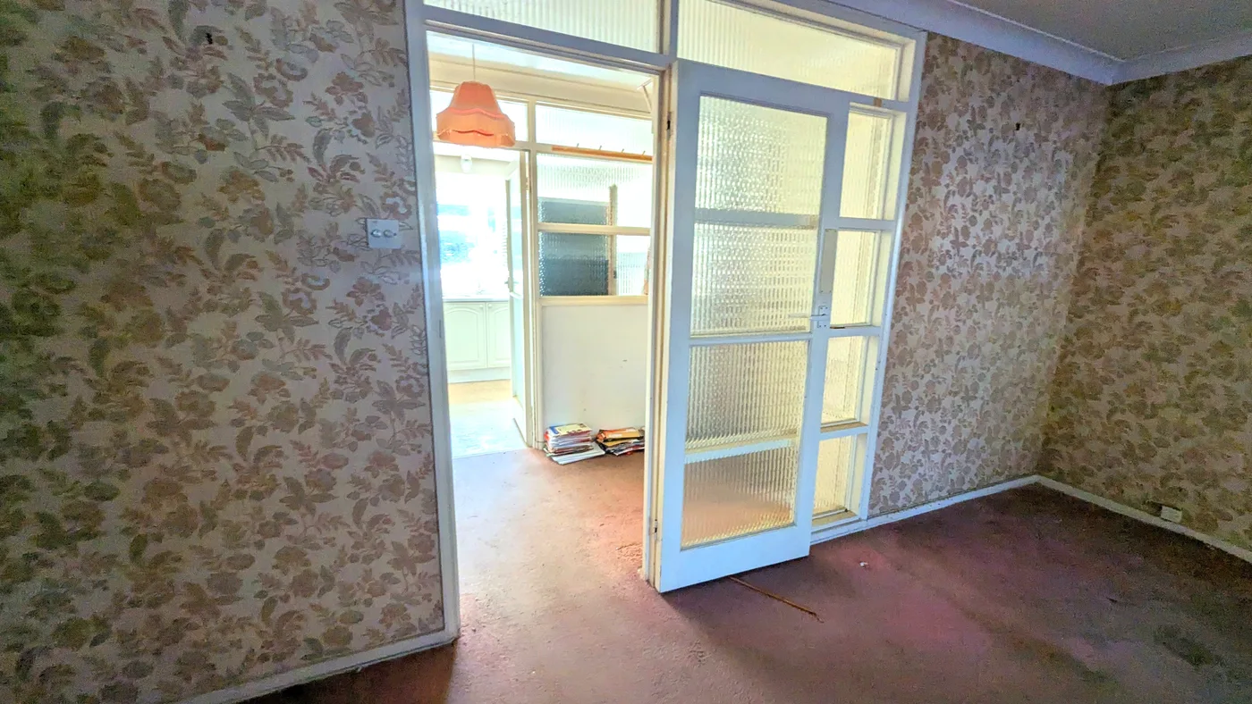 An empty room with dated floral wallpaper, glazed internal doors and worn carpet