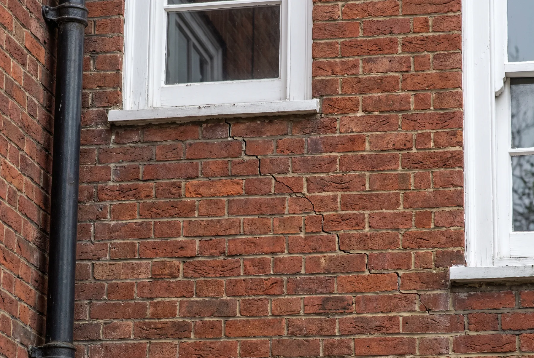 A fine stepped crack running diagonally through the red brickwork below a window of a 1930s block of flats, beside a drainpipe
